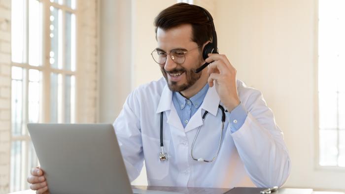Ein Arzt mit Headset und Stethoskop sitzt vor einem Laptop am Schreibtisch.